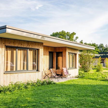 A small wooden house with a gray roof and a patio with chairs, located in a green landscape.