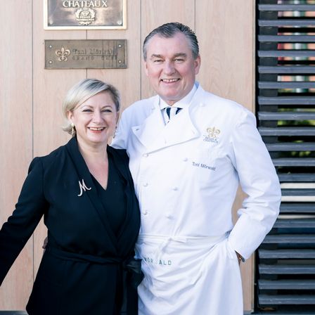 A smiling couple stands in front of a hotel with a sign reading 'Hotel & Restaurants' and a plaque labeled 'Relais & Chateaux'. The man in a chef's uniform smiles with his arm around the woman in a black robe.
