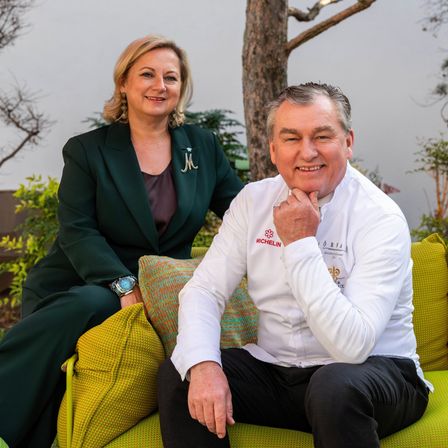 Ein Mann und eine Frau sitzen auf einer grünen Couch im Garten und lächeln in die Kamera. Der Mann trägt ein weißes Kochjackett mit Michelin-Marke.