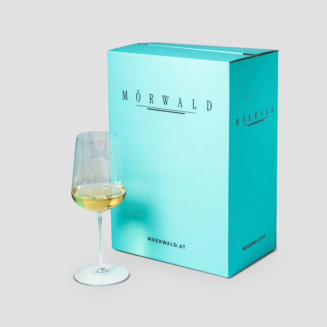 A turquoise box with the label MORWALD is placed on a white background. A wine glass with white wine is kept beside the box. The text MORWALD.AT is on the box.