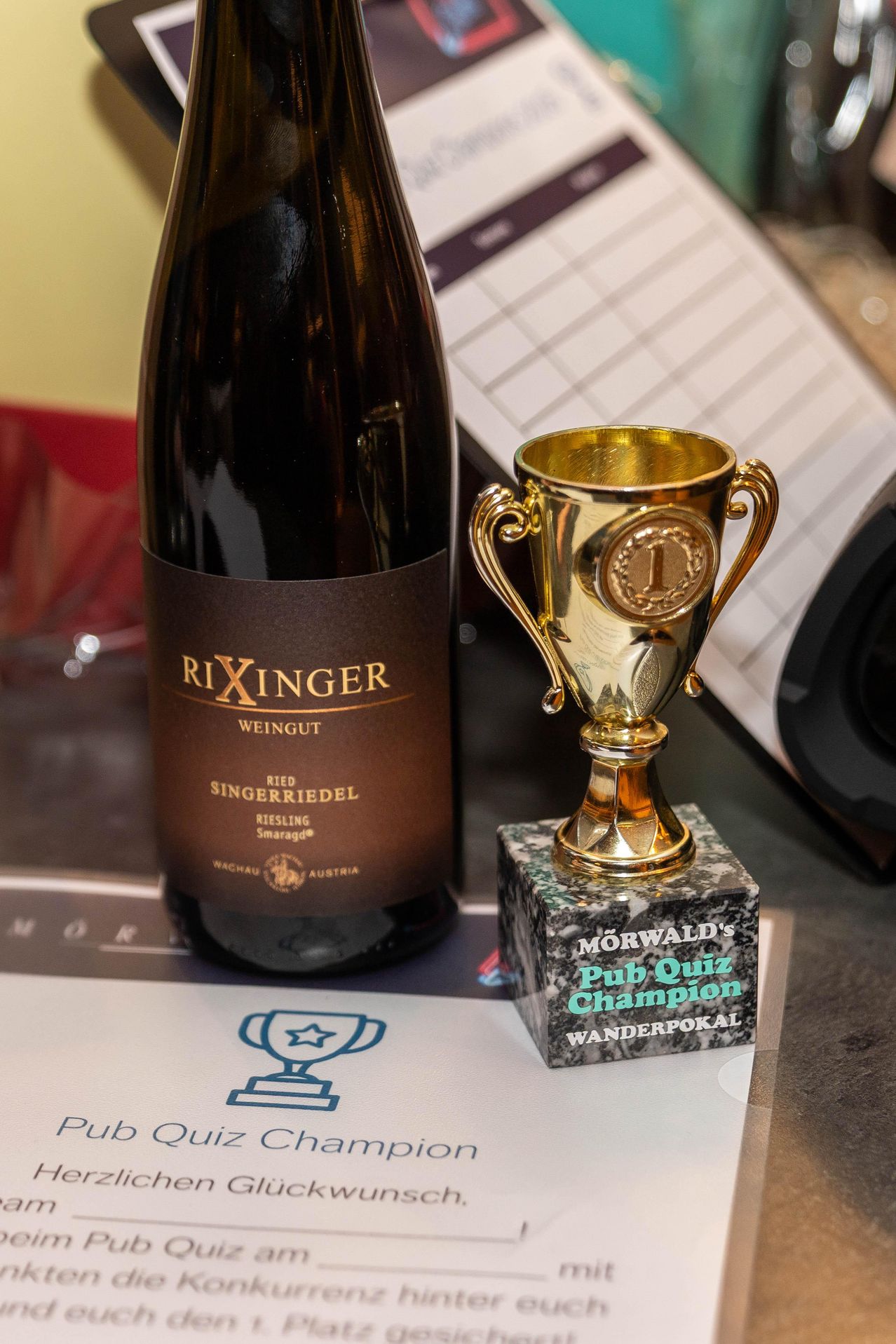 Eine Flasche Rixinger Wein und ein goldener Pokal mit der Aufschrift 'Morwald's Pub Quiz Champion' stehen auf einem Tisch. Der Pokal steht auf einem Granitsockel.