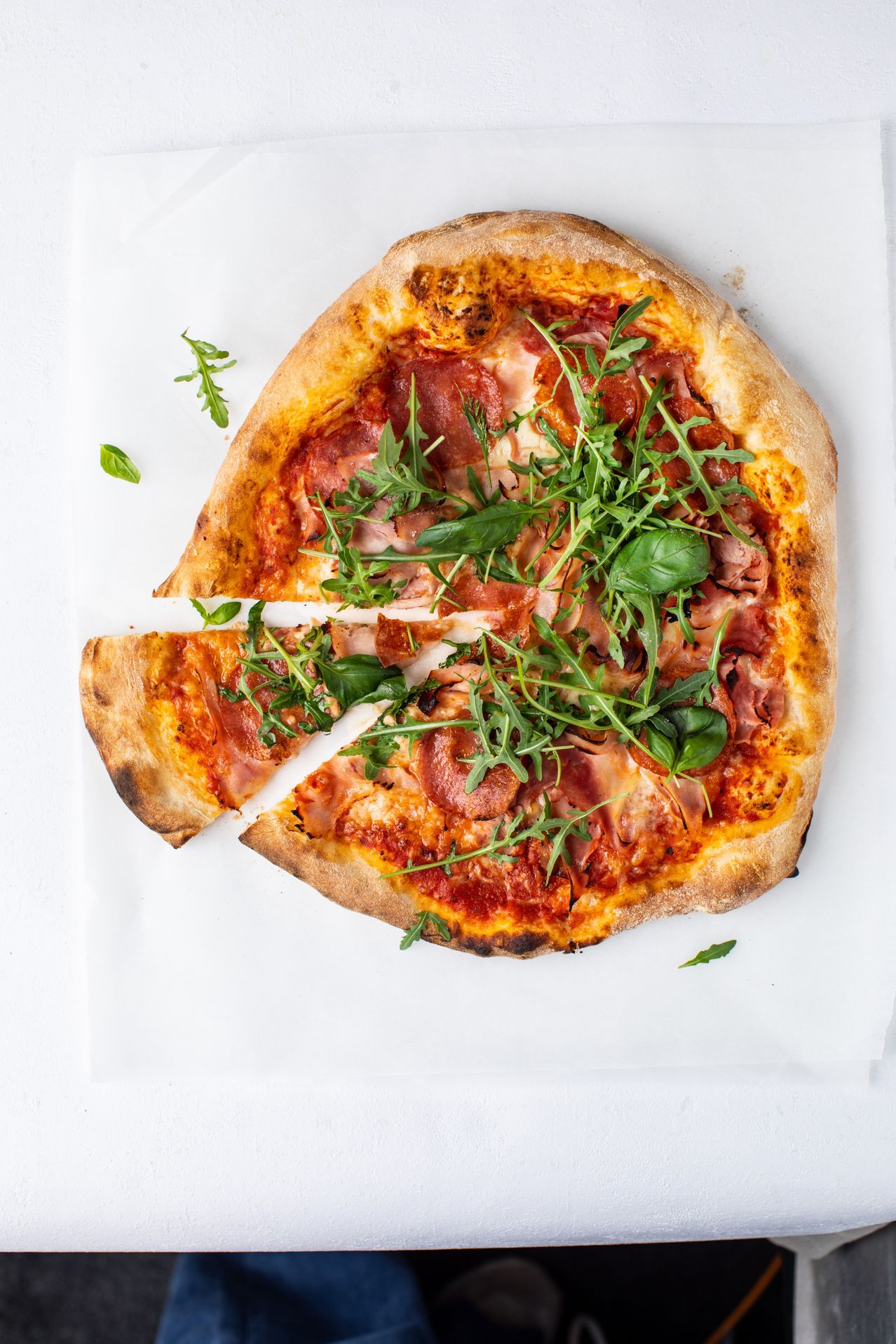 Eine ganze Pizza mit einem herausgenommenen Stück, belegt mit Schinken, Käse und Rucola auf weißem Hintergrund.