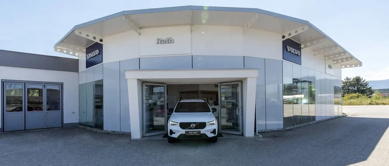 Ein weißer Volvo XC40 steht vor dem Volvo Rath-Händler, mit offenen Türen. Der Händler zeichnet sich durch ein modernes Design mit Glaswänden und einem blauen Schild aus.