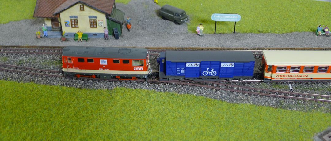 Ein Modellzugset mit einem blauen Fahrradwagen und einer roten Lokomotive, in der Nähe eines Hauses, eines Lieferwagens und eines Schildes.