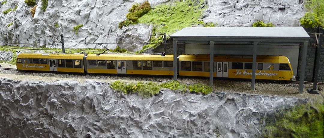Ein gelber Modellzug steht neben einem Bahnhof mit Dach auf einer Schiene. Der Bahnhof ist von einem moosbedeckten Felsvorsprung umgeben.