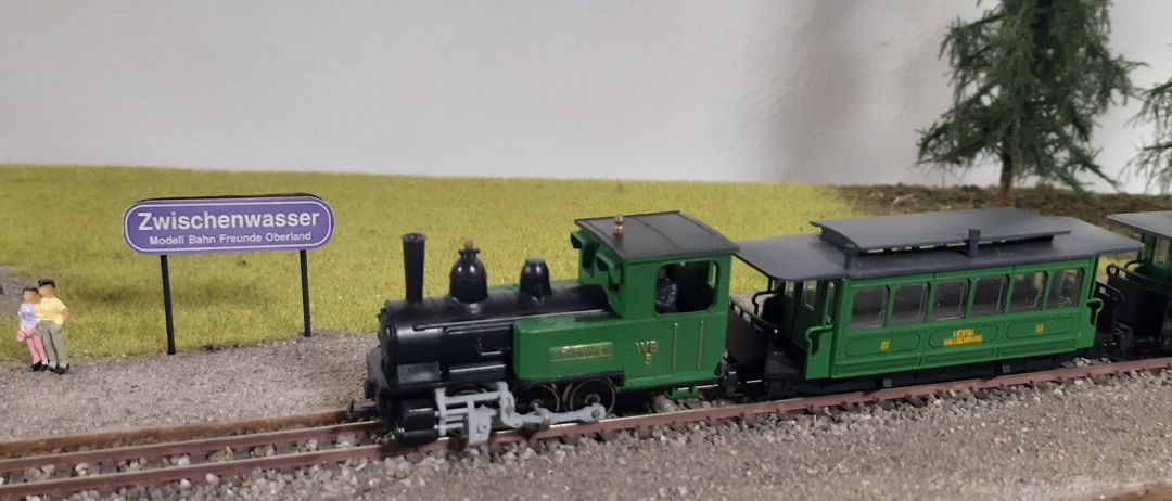 Eine grüne und schwarze Modelleisenbahn auf den Gleisen, mit einer Person im Inneren. Hinter dem Zug befindet sich ein Grasbereich mit einem Schild.