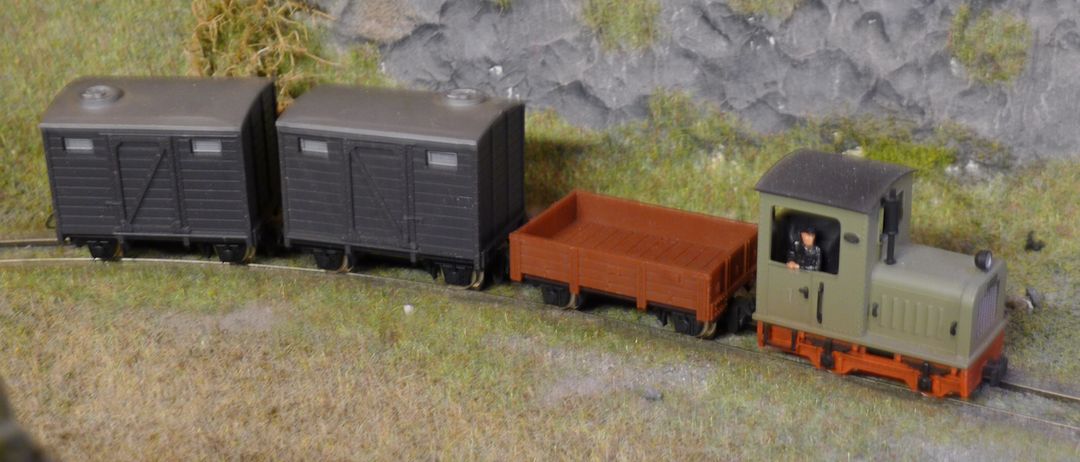 Ein Modellzug mit einem schwarzen Güterwagen und einem braunen Flachbettwagen auf einer Strecke mit Gras und einer Felswand im Hintergrund.