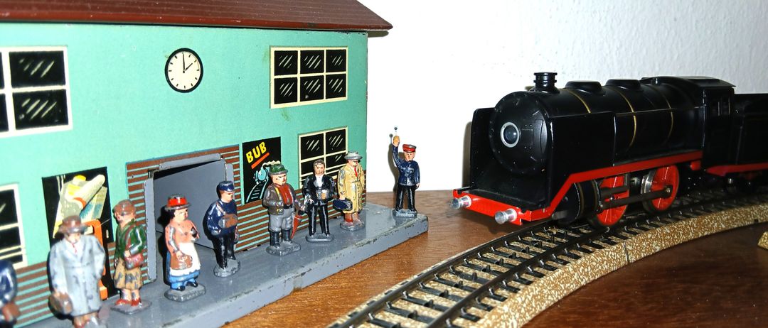 Eine Miniatur-Modelleisenbahnstation mit mehreren Menschen, die herumstehen, und einem Zug auf den Gleisen. Die Station hat eine Uhr und ein Schild mit der Aufschrift 'BUB'.