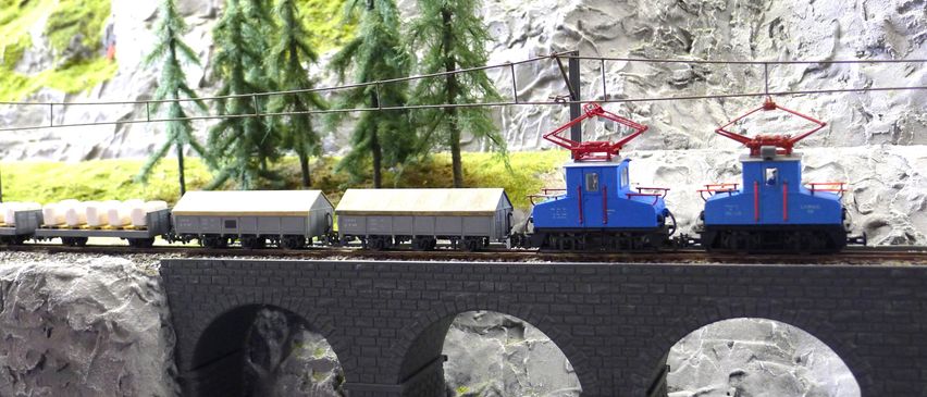 Ein Modellzug mit einer blauen Lokomotive und zwei grauen Waggons fährt über eine steinerne Bogenbrücke. Im Hintergrund befinden sich Bäume und eine felsige Klippe.