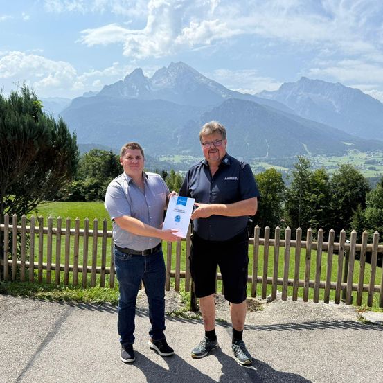 Zwei Männer stehen auf einer Straße und halten ein Zertifikat vor einem Holzzaun mit Blick auf einen Berg im Hintergrund.