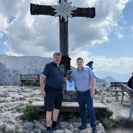 Zwei Männer stehen vor einem hölzernen Kreuz auf einem Berggipfel und halten ein Zertifikat. Hinter ihnen sind hölzerne Bänke und eine entfernte Bergkette unter einem teilweise bewölkten Himmel.