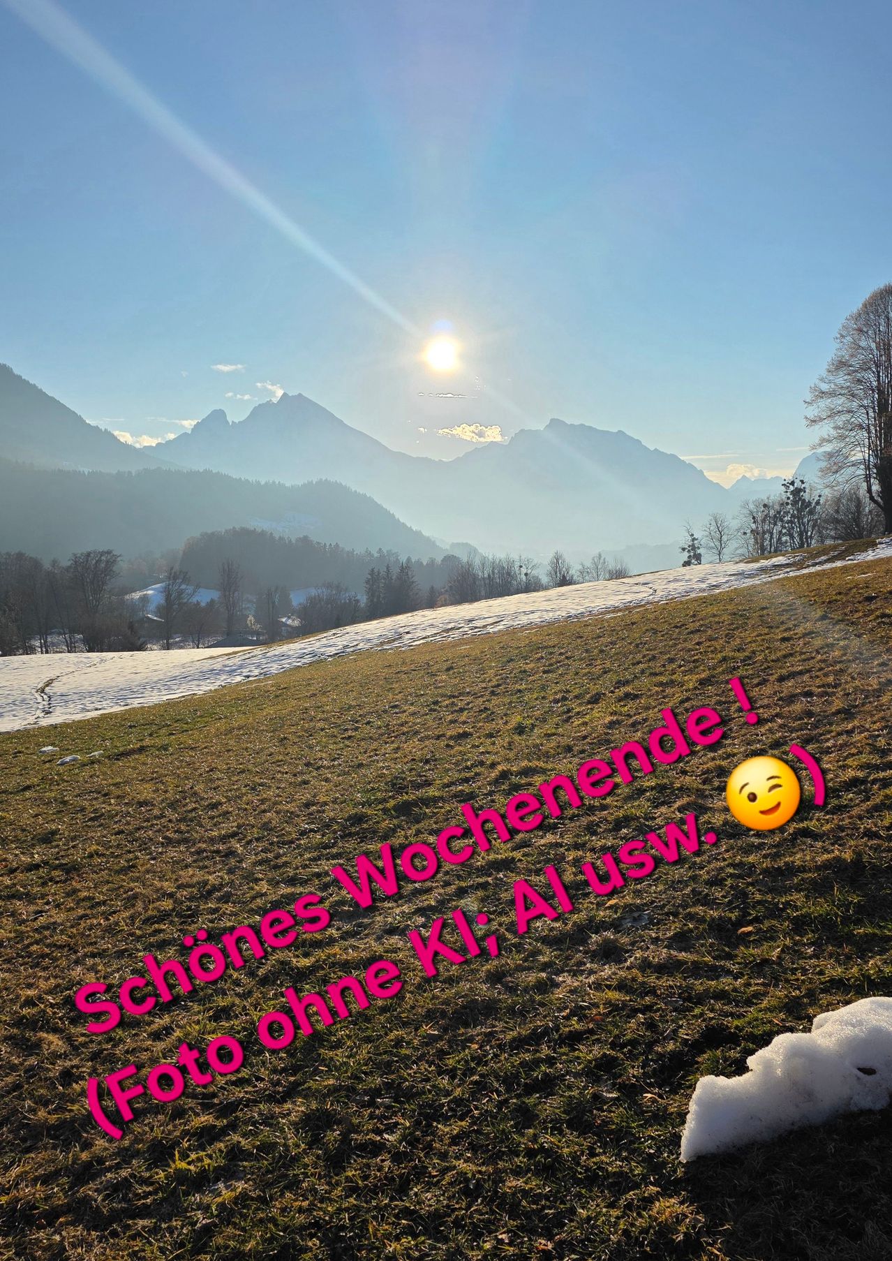 Ein sonniger Tag in einer verschneiten Landschaft mit Bergen im Hintergrund. Der Text in Pink lautet 'Schönes Wochenende!' und ein Smiley-Gesicht ist vorhanden.