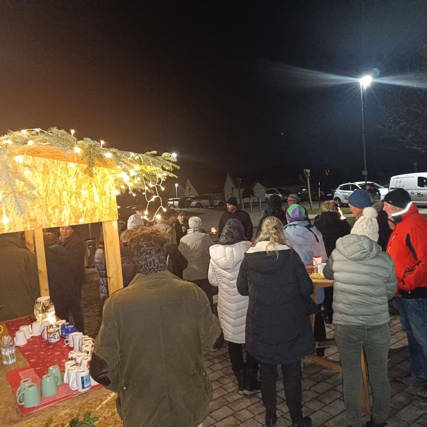 Eine Gruppe von Menschen steht bei Nacht um einen Tisch unter einem geschmückten Pavillon, einige halten Tassen. Der Bereich ist von einer Straßenlaterne beleuchtet.