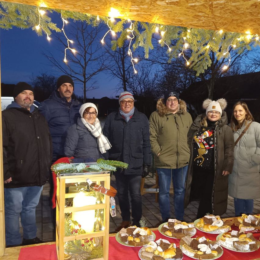 Eine Gruppe von Menschen in Winterkleidung steht unter einem Baldachin mit Lichtern. Sie lächeln und posieren für ein Foto. Ein Tisch vor ihnen hat Teller mit Desserts.