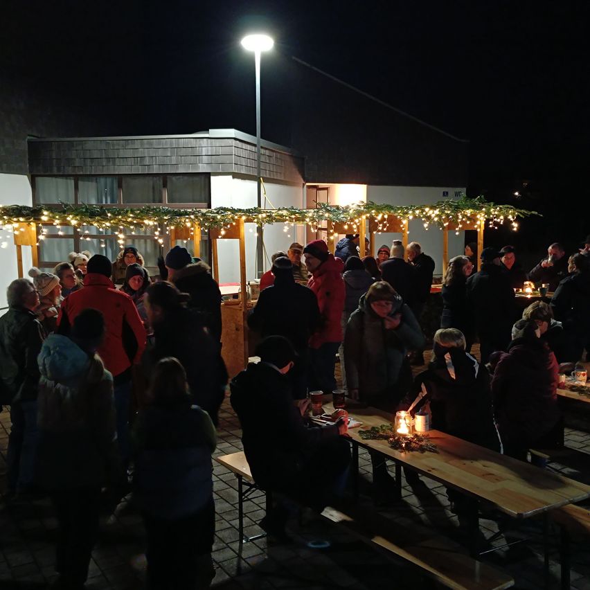 Eine Gruppe von Menschen, die sich warm gekleidet haben, versammelt sich nachts vor einem Gebäude. Sie sind von Tischen mit brennenden Kerzen und Essen umgeben. Das Gebäude hat Lichterketten, die am Dach hängen.