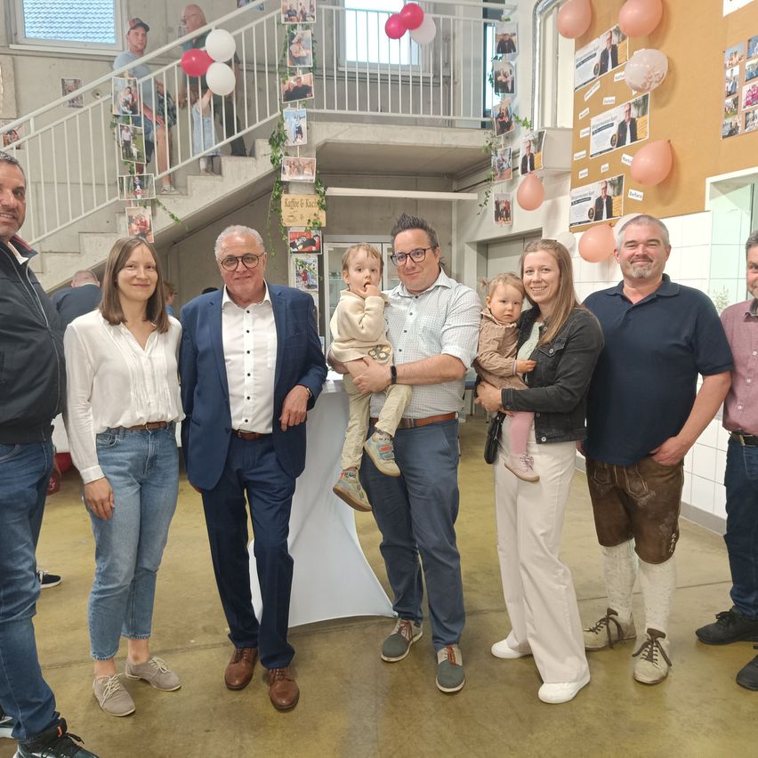 Eine Gruppe von Menschen posiert für ein Foto mit zwei Babys in einem Raum mit Luftballons und einer Treppe im Hintergrund.