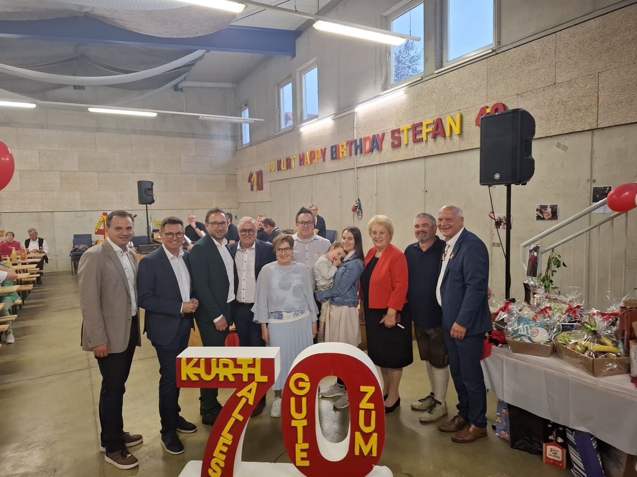 Eine Gruppe von Menschen steht hinter einer Geburtstagsdekoration mit einer Nummer 70 im Vordergrund. Im Hintergrund befindet sich ein Schild mit der Aufschrift 'Happy Birthday Stefan 40'. Ein Tisch mit Geschenken steht rechts.