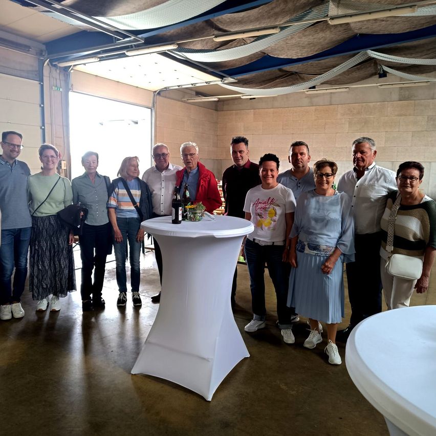 Eine Gruppe von Menschen, darunter Erwachsene und Senioren, posiert für ein Foto in einem Raum mit weißen Tischen und Flaschen.