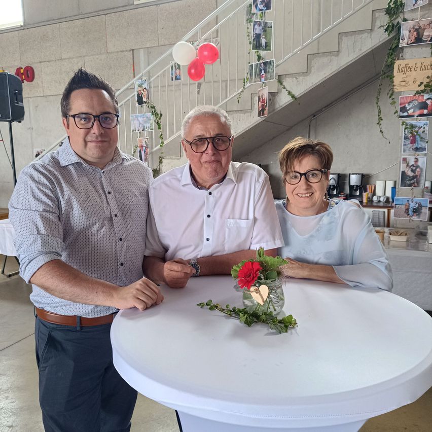 Drei Personen, zwei Männer und eine Frau, stehen um einen Tisch mit einem Blumenarrangement. Der Mann links trägt einen Gürtel und eine Brille, während der Mann in der Mitte eine Uhr trägt. Die Frau rechts lächelt und trägt eine Brille.