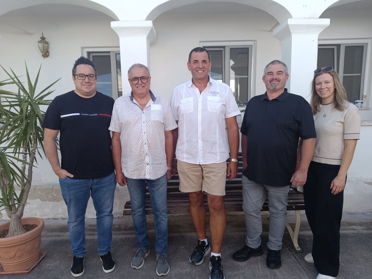 Five men stand in front of a white building, smiling and posing for a photo. The man in the middle wears a white shirt and shorts. The man on the left wears a black shirt and glasses. The man on the right wears a black polo shirt.