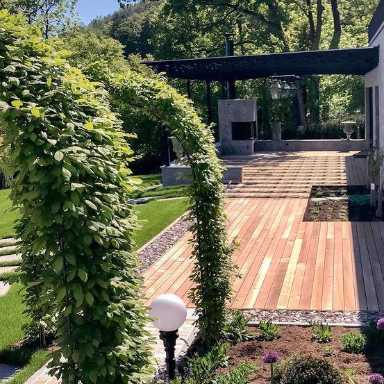 Ein Garten mit einer Holzterrasse, umgeben von Pflanzen und Bäumen. Es gibt einen Kamin, eine Überdachung und einen Rasenbereich mit einer weißen Lampe.