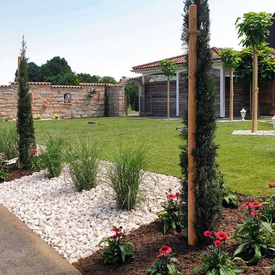 Ein Hinterhof mit Rasen, weißem Kies, Pflanzen und Bäumen, einer Steinmauer und einem Haus mit einem Ziegeldach im Hintergrund.