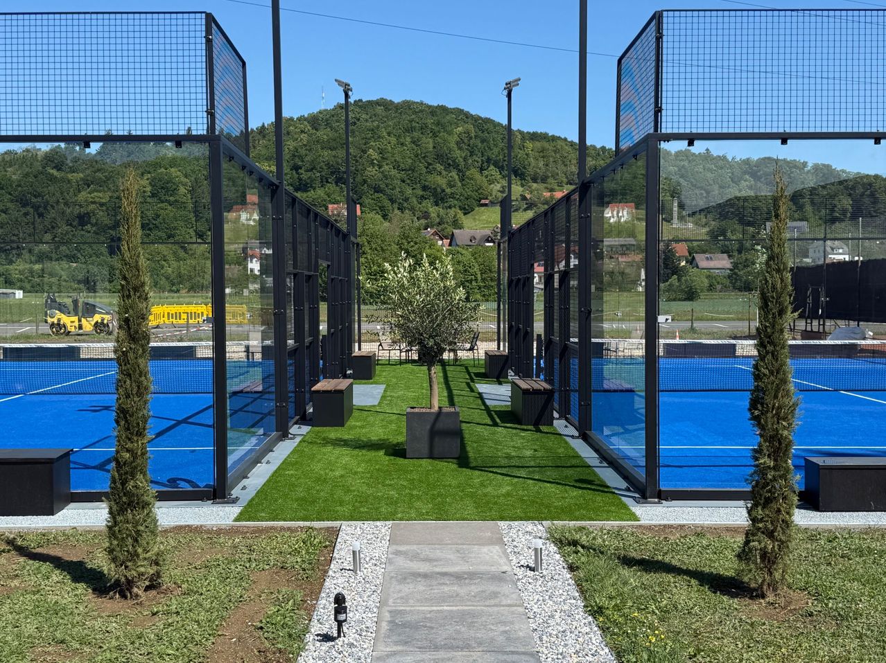 Ein Tennisplatz mit blauem Boden, umgeben von Metallzäunen und einem Rasen. Bänke und ein Topf mit Pflanzen stehen in der Mitte. Ein gelber Zug ist in der Ferne zu sehen.