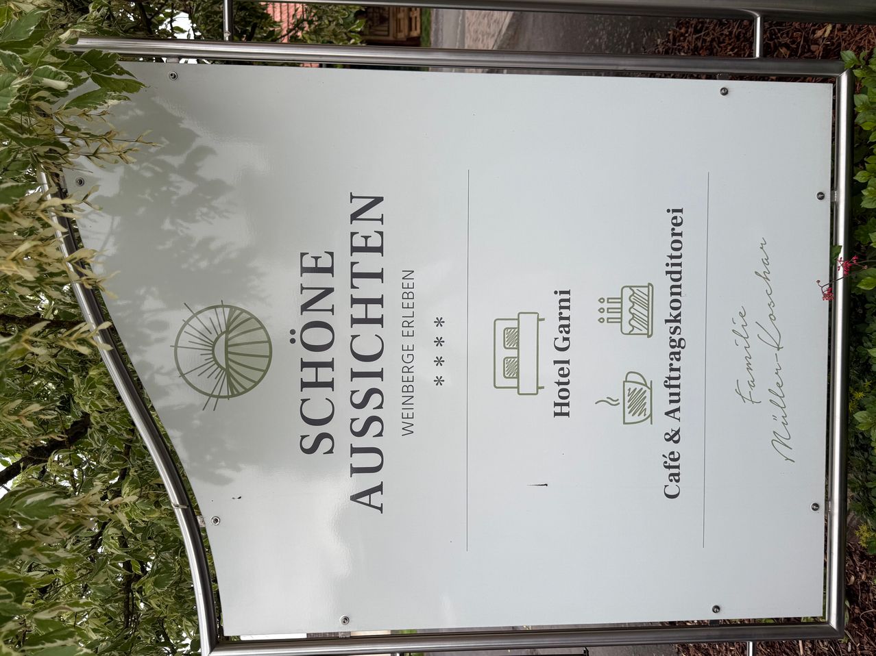 A white sign advertises 'Schon Aussichten' with icons for Hotel Garni, a cafe, and a coffee machine. The image is set in a landscape with green leaves and a metal structure.