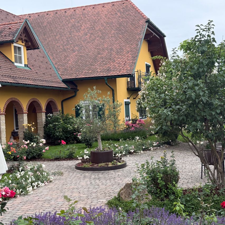 A yellow house with a red roof and a garden with various flowers and plants. The house has arched windows and a balcony.
