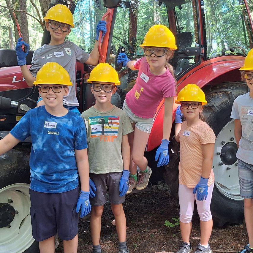 Sechs Kinder, alle mit Schutzhelmen, posieren neben einem roten Traktor. Die Kinder tragen verschiedene T-Shirts, einige mit Logos, und alle tragen blaue Handschuhe. Bäume umgeben die Szene.