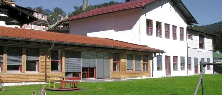 Ein modernes Gebäude mit rotem Dach und weißen Wänden steht in einem bergigen Gebiet. Das Gebäude hat mehrere Fenster mit roten Rahmen. Vor dem Gebäude befindet sich ein Spielplatz mit einer Rutsche und einer Schaukel. Vor dem Gebäude liegt eine Rasenfläche.