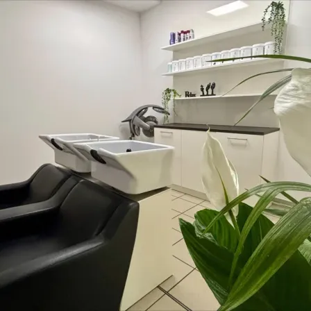 Ein leerer Salon mit weißen Wänden, einer weißen Arbeitsplatte, Regalen mit Produkten, drei Waschbecken und einem schwarzen Ledersessel.