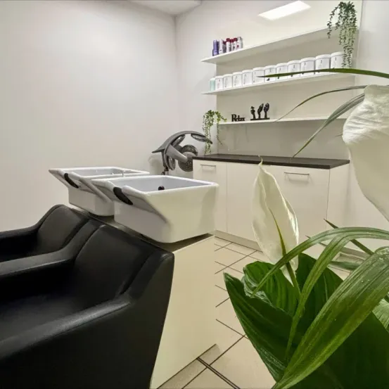 Ein leerer Salon mit weißen Wänden, einer weißen Arbeitsplatte, Regalen mit Produkten, drei Waschbecken und einem schwarzen Ledersessel.