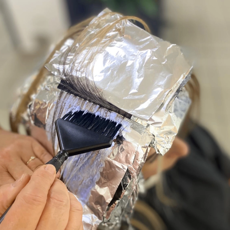 Eine Hand hält einen Pinsel und trägt Farbe auf das mit Folie bedeckte Haar auf. Die Hand der Person ist sichtbar, ebenso wie ein Ring. Die Folie ist ordentlich um den Kopf gewickelt.