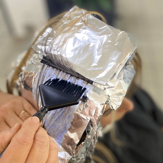 Eine Hand hält einen Pinsel und trägt Farbe auf das mit Folie bedeckte Haar auf. Die Hand der Person ist sichtbar, ebenso wie ein Ring. Die Folie ist ordentlich um den Kopf gewickelt.