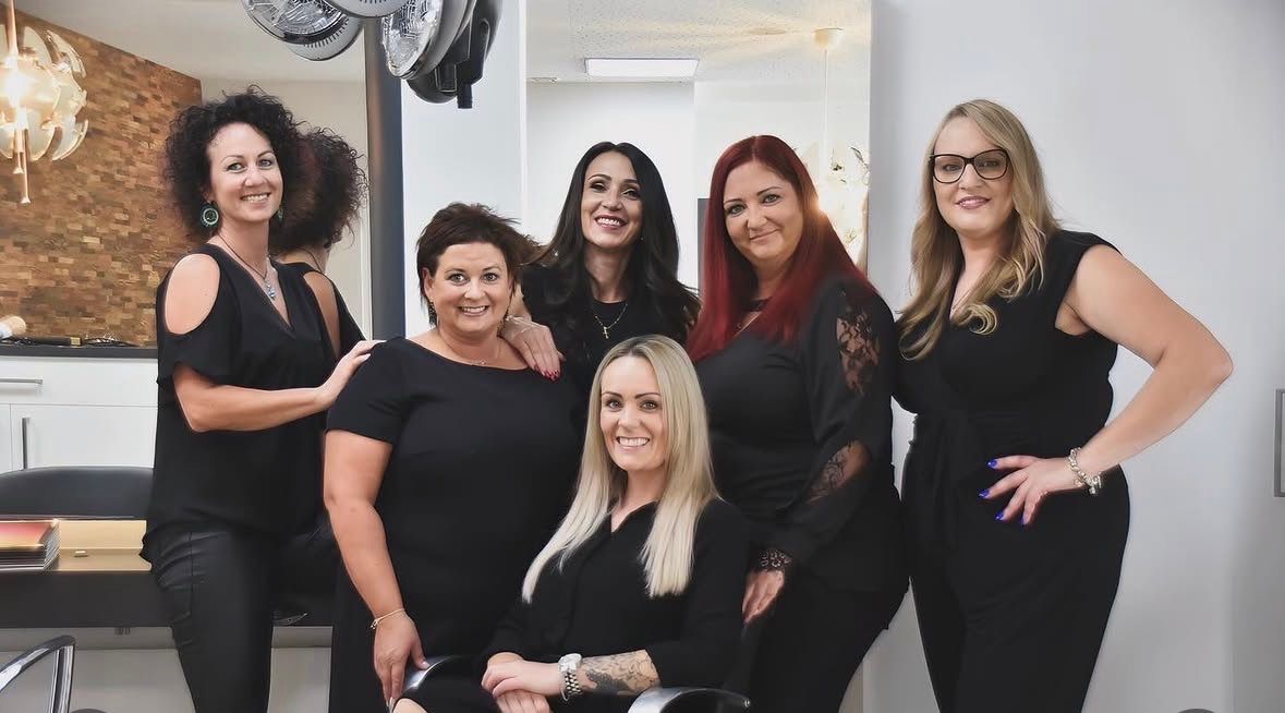 A group of women in black dresses are posing for a photo in a salon. They are smiling and standing close to each other. The woman in the middle is sitting on a chair. Behind them is a mirror with a reflection of a lamp and a ceiling fan.