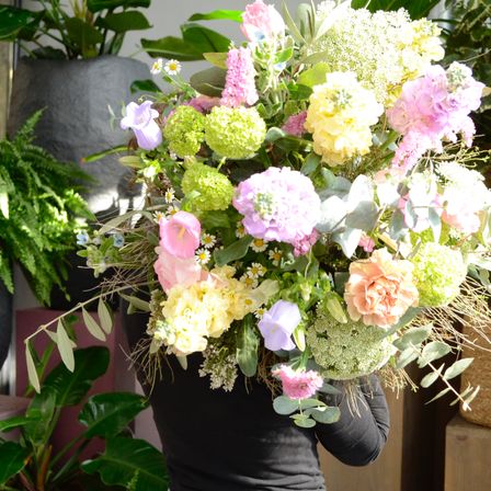 Eine Person trägt einen großen Blumenstrauß mit verschiedenen Blumenfarben und grünen Blättern, umgeben von Topfpflanzen.