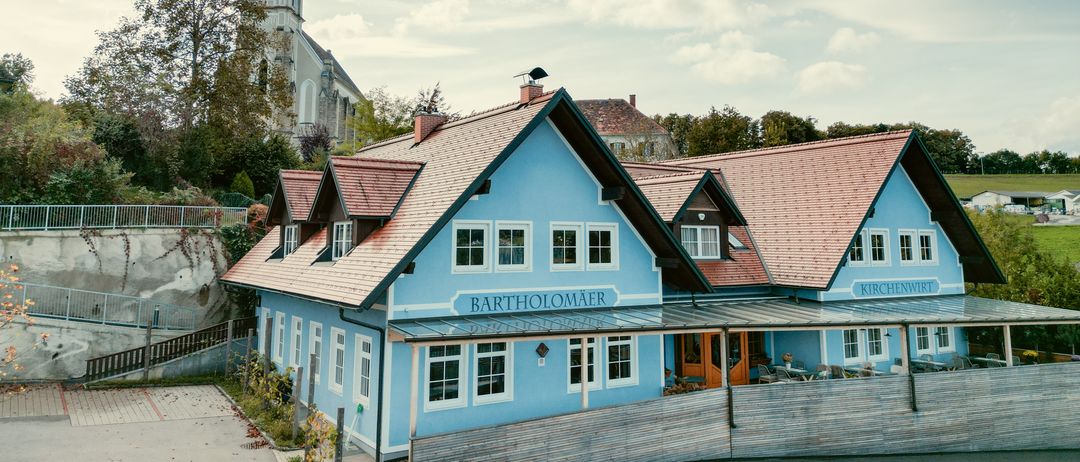 Ein blaues Gebäude namens BARTHOLOMAER mit einem roten Dach steht vor einer Kirche unter einem bewölkten Himmel.