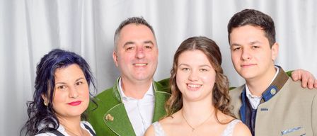 Eine lächelnde Familie mit vier Personen posiert zusammen. Die Frau auf der linken Seite trägt ein traditionelles Kleid, während der Mann neben ihr einen grünen Mantel trägt. Das Mädchen in der Mitte ist in einem weißen Kleid und der Mann auf der rechten Seite trägt einen braunen Anzugsmantel.