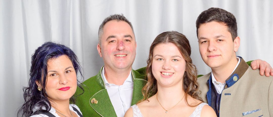 Eine lächelnde Familie mit vier Personen posiert zusammen. Die Frau auf der linken Seite trägt ein traditionelles Kleid, während der Mann neben ihr einen grünen Mantel trägt. Das Mädchen in der Mitte ist in einem weißen Kleid und der Mann auf der rechten Seite trägt einen braunen Anzugsmantel.