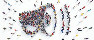 Eine große Menschenmenge ist in Herzform auf einer weißen Oberfläche versammelt. Sie tragen verschiedene Farben, und einige bewegen sich, während andere still stehen. Schatten werden auf die Oberfläche geworfen.