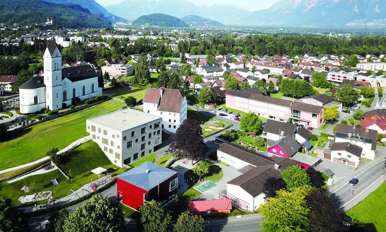 Ein Luftbild einer Stadt mit vielen Gebäuden, Bäumen und Bergen im Hintergrund. Es gibt eine weiße Kirche mit einem Turm und ein rotes Haus mit einem weißen Dach.