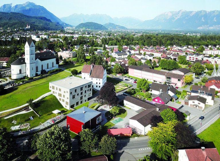 Ein Luftbild einer Stadt mit vielen Gebäuden, Bäumen und Bergen im Hintergrund. Es gibt eine weiße Kirche mit einem Turm und ein rotes Haus mit einem weißen Dach.