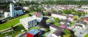 Ein Luftbild einer Stadt mit vielen Gebäuden, Bäumen und Bergen im Hintergrund. Es gibt eine weiße Kirche mit einem Turm und ein rotes Haus mit einem weißen Dach.