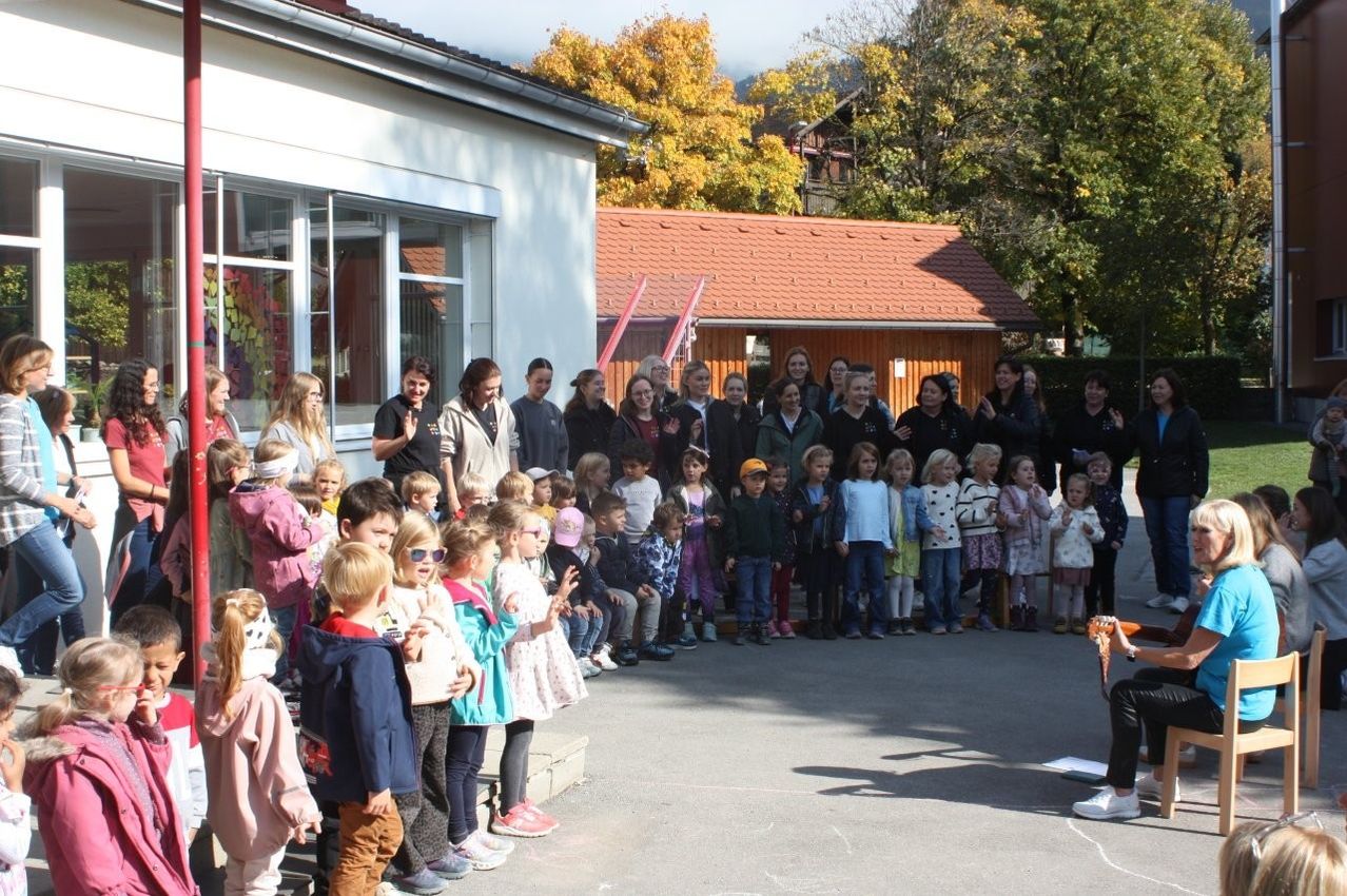 Eine Gruppe von Kindern und Erwachsenen versammelt sich sonnigen Tages vor einem Gebäude, mit Bäumen und Häusern im Hintergrund.