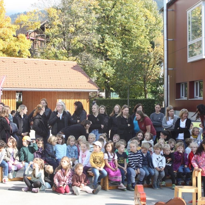 Eine Gruppe von Kindern und Erwachsenen versammelt sich vor einem Gebäude mit Bäumen und Bergen im Hintergrund. Einige sitzen, während andere dahinter stehen.