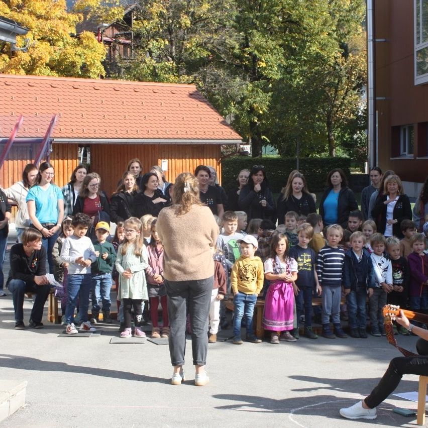 Eine Gruppe von Menschen, einschließlich Kindern und Erwachsenen, versammelt sich im Freien. Einige sitzen auf Bänken, andere stehen. Im Hintergrund befinden sich Bäume und ein Gebäude.