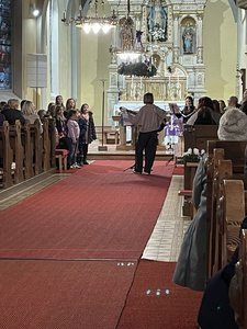Ein Chor singt in einer Kirche. Ein erwachsener Mann steht in der Mitte und hält ein Notenpult. Kinder und Erwachsene stehen um ihn herum.