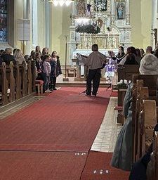 Ein Chor singt in einer Kirche. Ein erwachsener Mann steht in der Mitte und hält ein Notenpult. Kinder und Erwachsene stehen um ihn herum.