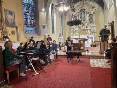 Eine Gruppe von Kindern und Erwachsenen sitzt auf einem roten Teppich in einer Kirche. Ein Mann steht hinter einem Podium, und ein anderer spielt Klavier. Ein Priester ist am Altar mit einer Blumenarrangement.